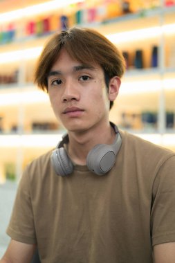 Kahverengi tişörtlü genç Taylandlı öğrenci sosyal medya, uzaktan iletişim ve dijital yaşam tarzı gibi modern bir kafede boynuna kulaklık takıyor..