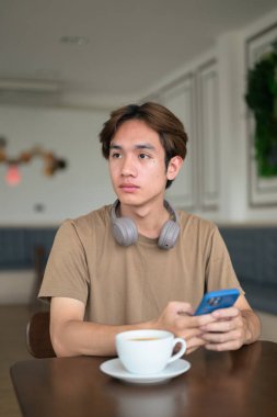Kahverengi tişörtlü genç Taylandlı öğrenci sosyal medya, uzaktan iletişim ve dijital yaşam tarzı gibi modern bir kafede boynuna kulaklık takıyor..
