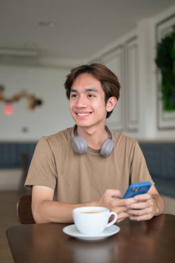 Kahverengi tişörtlü genç Taylandlı öğrenci sosyal medya, uzaktan iletişim ve dijital yaşam tarzı gibi modern bir kafede boynuna kulaklık takıyor..