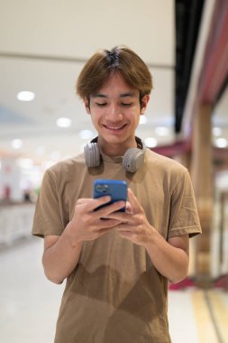 20 yaşında boynunda kulaklıkla Taylandlı bir adam alışveriş merkezinin koridorunda telefonuna gülümserken, mesajlaşırken, online alışveriş yaparken ve şehir yaşamında.