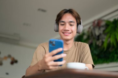 Kahverengi tişörtlü genç Taylandlı öğrenci sosyal medya, uzaktan iletişim ve dijital yaşam tarzı gibi modern bir kafede boynuna kulaklık takıyor..