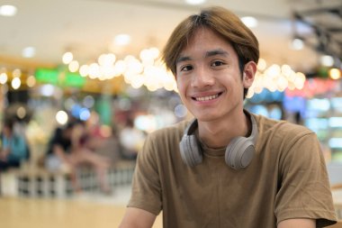 Kahverengi tişörtlü genç Taylandlı öğrenci sosyal medya, uzaktan iletişim ve dijital yaşam tarzı gibi modern bir kafede boynuna kulaklık takıyor..