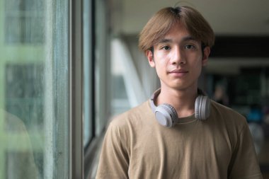 Genç yakışıklı ve zayıf Asyalı Taylandlı adam spor salonunda pencerenin yanında kulaklık takıyor.