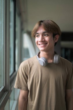 Genç yakışıklı ve zayıf Asyalı Taylandlı adam spor salonunda pencerenin yanında kulaklık takıyor.
