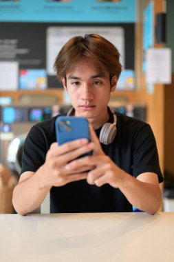 Genç yakışıklı Taylandlı adam çamaşır makinesinde oturmuş telefon kullanıyor, siyah tişört giyiyor. Gen Z yaşam tarzını, günlük ev işlerini, çamaşırları ve şehir yaşamını gösteren samimi bir portre..