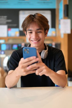 Genç yakışıklı Taylandlı adam çamaşır makinesinde oturmuş telefon kullanıyor, siyah tişört giyiyor. Gen Z yaşam tarzını, günlük ev işlerini, çamaşırları ve şehir yaşamını gösteren samimi bir portre..