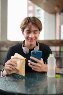 Mutlu Asyalı genç Taylandlı öğrenci dışarıda öğle yemeği için sandviçini yerken oturup telefonla konuşuyor.