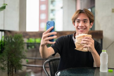 Mutlu Asyalı genç Taylandlı öğrenci dışarıda öğle yemeği için sandviçini yerken oturup telefonla konuşuyor.