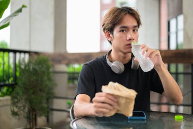 Mutlu Asyalı genç Taylandlı öğrenci sandviç yiyor ve öğle yemeği için açık havada su içiyor.