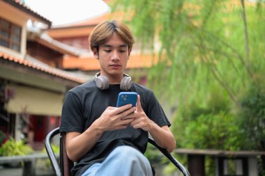 Genç Taylandlı bir adam geleneksel Çin bahçe pazarında akıllı telefon kullanarak dışarıda oturuyor. Çeşitli kavramlar ve kullanımlar için kendine güvenen, rahat ve otantik görünüm