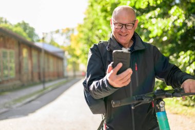 Portrait of a smiling senior man riding a electric kick scooter in city and using smartphone
