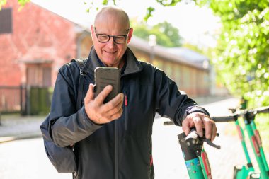 Portrait of a smiling senior man riding a electric kick scooter in city and using smartphone