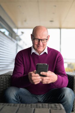 Portrait of a happy senior Scandinavian man using a mobile phone while sitting in an airport armchair