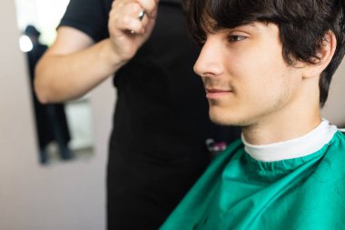 Salonda genç bir adam var. Kuaför, makas ve tarakla şık bir saç kesimi yapar.
