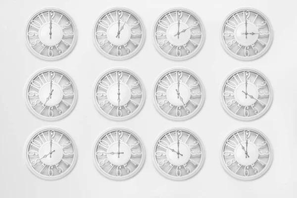 12 circle white modern clocks in a row by 4 on white background wall. The time on the clock varies from 0 to 12 o'clock