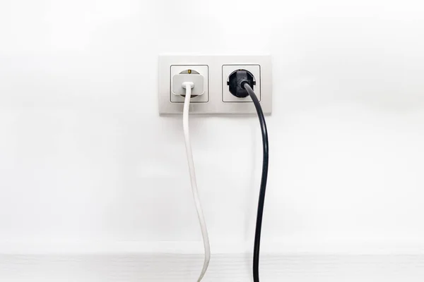 White double outlet installed on the white wall with inserted phone adapter and a black electrical plug, front view.