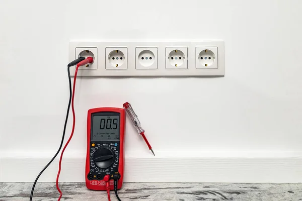 White five-way wall power socket installed on the white wall. Multimeter measures voltage