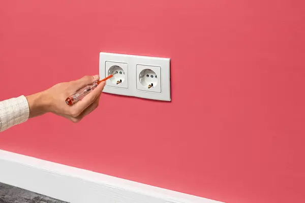 Human hand is holding a screwdriver plugged into an outlet. White double socket installed on the pink wall, side view.