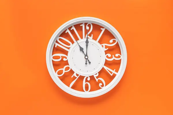 Modern white clock with a circle on a orange wall background, eleven o\'clock on the clock. Elegant clock, combination of white and orange colors