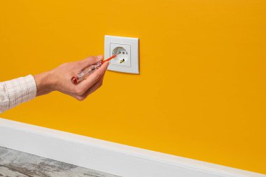İnsan eli prize takılı bir tornavida tutuyor. Sarı duvara yerleştirilmiş beyaz soket, yan görünüm.