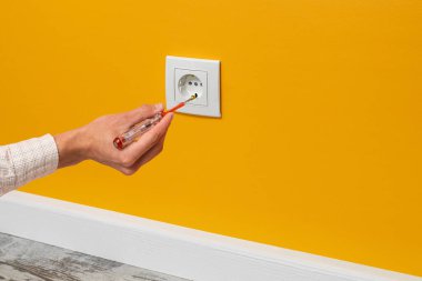 İnsan eli prize takılı bir tornavida tutuyor. Sarı duvara yerleştirilmiş beyaz soket, yan görünüm.