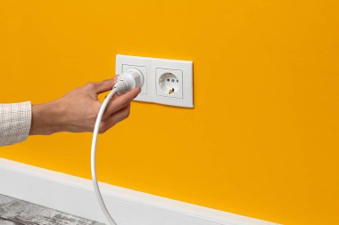 İnsan eli, sarı duvardaki çift beyaz elektrik prizine bağlı beyaz kabloyu tutuyor.