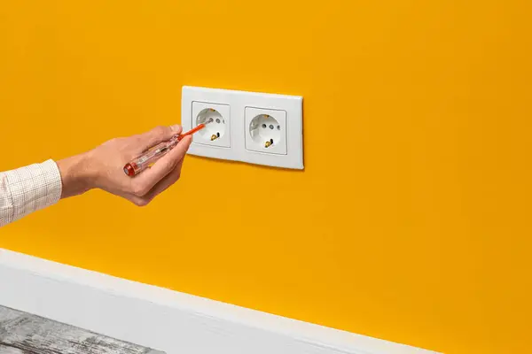 Human hand is holding a screwdriver plugged into an outlet. White double socket installed on the yellow wall, side view.