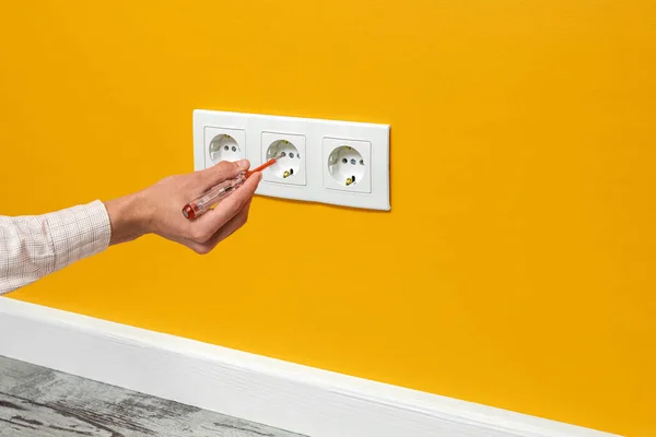 Human hand is holding a screwdriver plugged into an outlet. White triple socket installed on the yellow wall, side view.