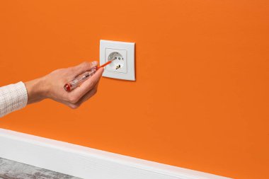 İnsan eli prize takılı bir tornavida tutuyor. Turuncu duvara yerleştirilmiş beyaz soket, yan görünüm.