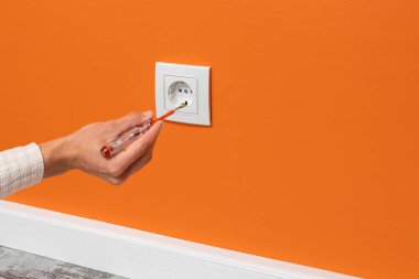 İnsan eli prize takılı bir tornavida tutuyor. Turuncu duvara yerleştirilmiş beyaz soket, yan görünüm.