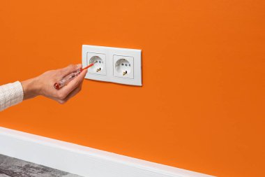 İnsan eli prize takılı bir tornavida tutuyor. Turuncu duvarda beyaz çift soket, yan görüş.