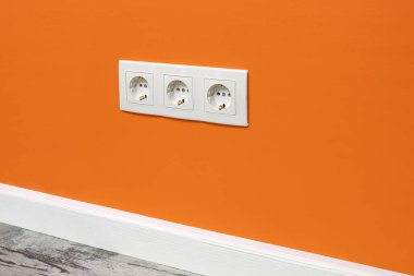 Turuncu duvarda beyaz üçlü priz, yan görünüm.