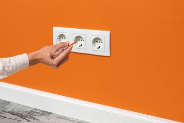 İnsan eli prize takılı bir tornavida tutuyor. Turuncu duvara yerleştirilmiş beyaz üçlü soket, yan görüş.