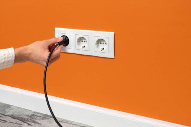 İnsan eli, turuncu duvardaki üçlü beyaz elektrik prizinden siyah kabloyu çekiyor.