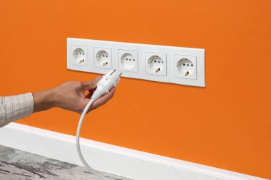 İnsan eli, turuncu duvardaki beş yönlü bir prize beyaz kablo takıyor.