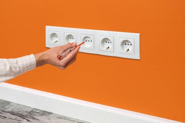 İnsan eli prize takılı bir tornavida tutuyor. Turuncu duvara yerleştirilmiş beyaz beş kanallı güç prizi..