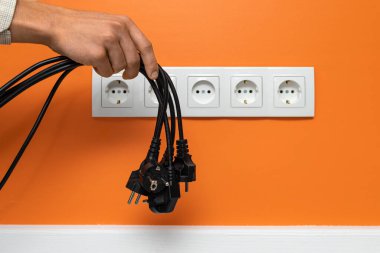 Turuncu duvardaki beş yönlü bir prizin önünde siyah kabloyu tutan insan eli. Elektrik prizleri avuçtan nazikçe sarkıyor