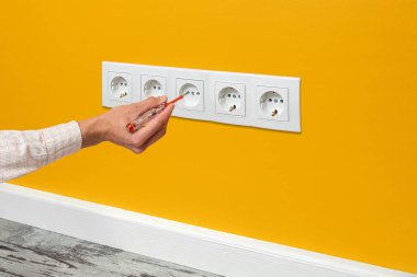 İnsan eli prize takılı bir tornavida tutuyor. Sarı duvara, yan görüşe yerleştirilmiş beş kanallı beyaz priz..