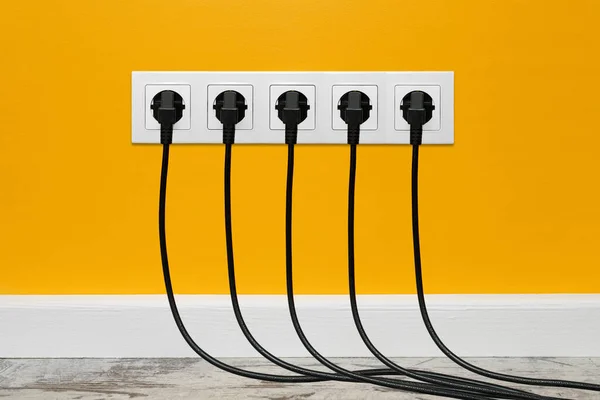 White five-way wall power socket installed on a yellow wall with five black plugs inserted. The outlet is filled with black cords, front view.