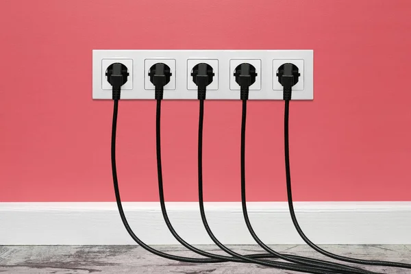 White five-way wall power socket installed on a pink wall with five black plugs inserted. The outlet is filled with black cords, front view.