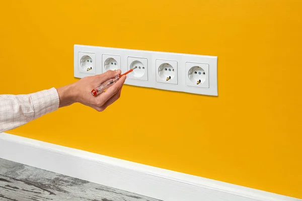 Human hand is holding a screwdriver plugged into an outlet. White five-way wall power socket installed on the yellow wall, side view.