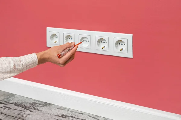 Human hand is holding a screwdriver plugged into an outlet. White five-way wall power socket installed on the pink wall, side view.