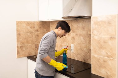 Genç bir adam mutfak masasını temizleme spreyi, lastik eldiven ve bez kullanarak temizliyor.