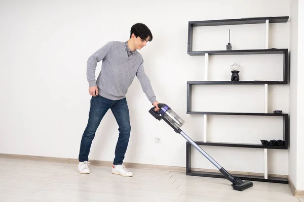 A young man is meticulously cleaning a laminate floor in a white room with a cordless vacuum cleaner, showing pleasure in his chores. In the background, there's a modern bookshelf with decorative objects.