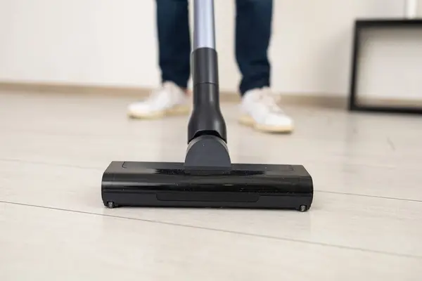 Close-up: A young man is using a cordless vacuum cleaner to clean a laminate floor in a white room. The blurred background focuses only on the cleaning action.