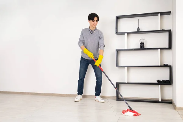 A young man wearing yellow household gloves is mopping a laminate floor in a white room. In the background, there\'s a stylish, ornate bookcase.