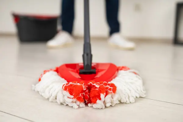 Close-up of the mop head on the laminate floor, held by a man standing near a bucket with a blurred background in a white room.