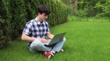 Şehir parkında yeşil çimlerin üzerinde oturan genç bir adam dizüstü bilgisayarla çalışıyor..