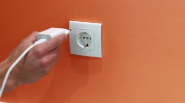 Bir el, bir telefon şarjını turuncu bir duvardaki beyaz bir elektrik prizine takıyor..