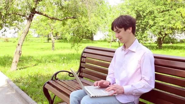 一位年轻人坐在公园的长椅上 在笔记本电脑上工作 周围环绕着生机勃勃的绿叶 营造出一种宁静自然的氛围 — 图库视频影像
